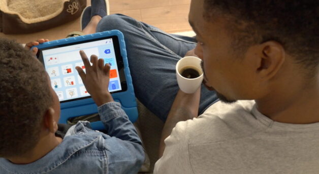 Son talking to father using simPODD on the couch, father holding a cup of coffee