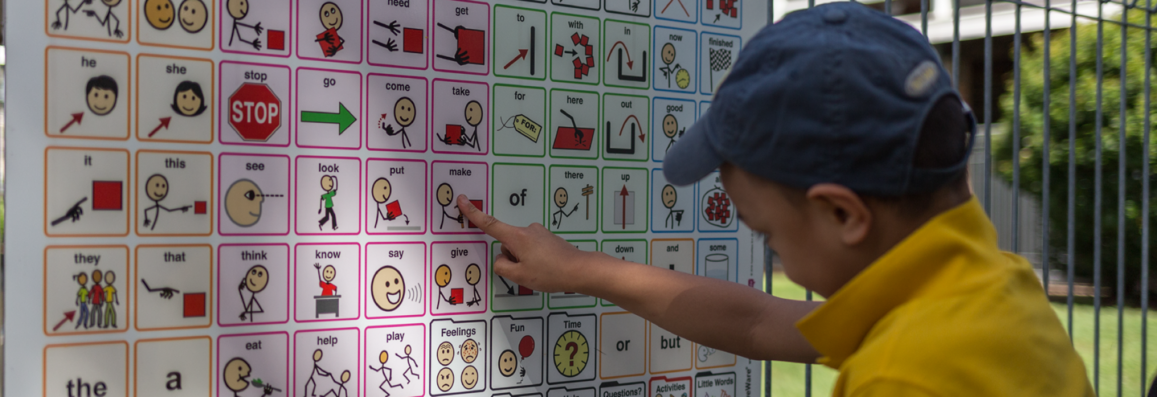 A boy with a cap on points at a printed core word board at the work and symbol for make