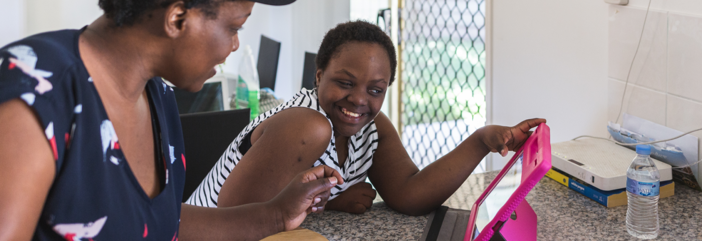 A parent sits with their child at a table. The child is smiling while touching their iPad.