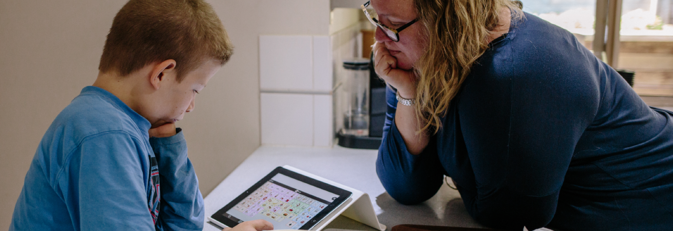 Een volwassene en een kind zitten tegenover elkaar. Het kind gebruikt een iPad met Proloquo2Go.