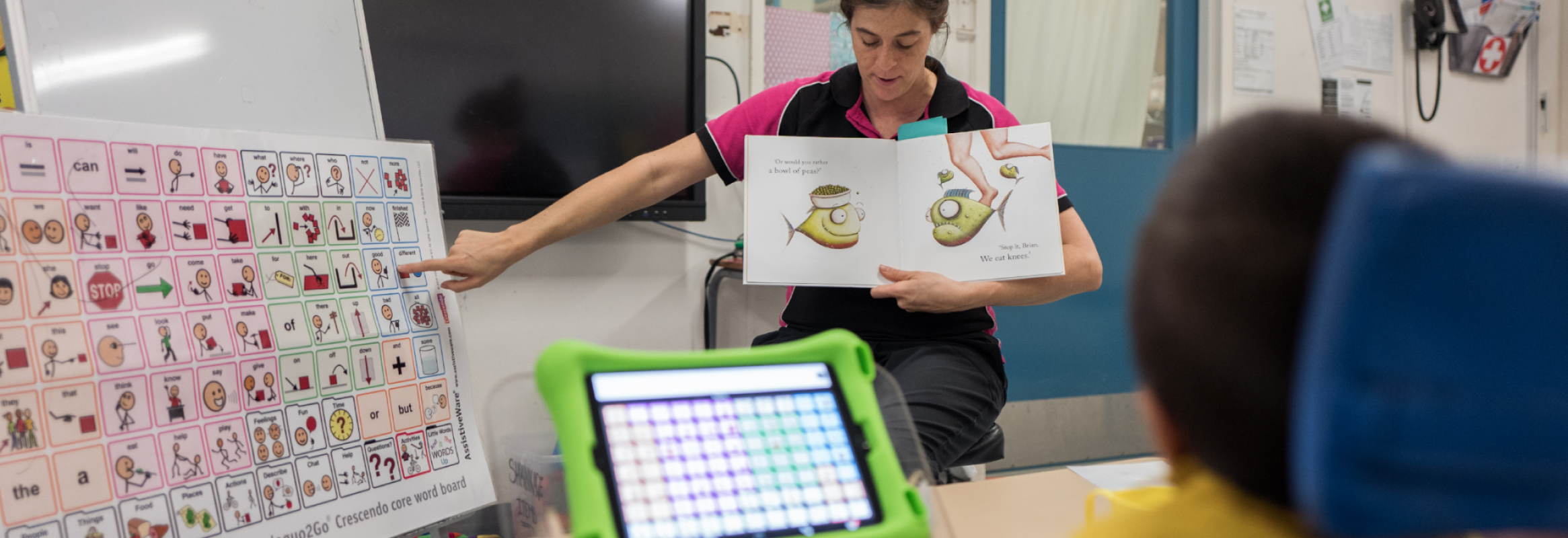 A teacher looking down a story book while pointing to a symbol on a large core word board.