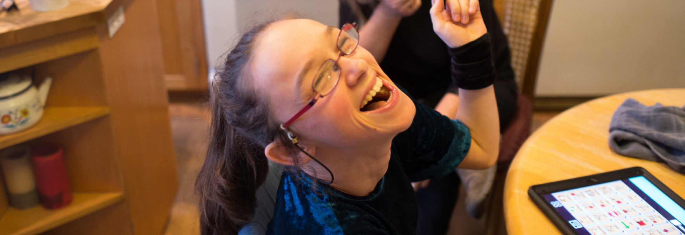 Girl smiles wide with her arm in the air. An iPad with Proloquo2Go is on the table infront of her.