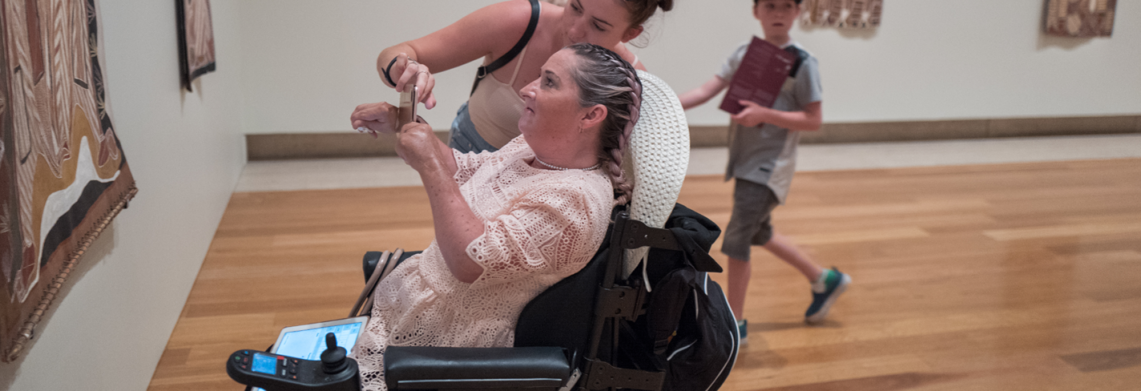 Two woman look at a quilt on a wall in a museum. One woman is in a wheelchair and is holding up her phone to take a picture. The other woman is standing and helping her.